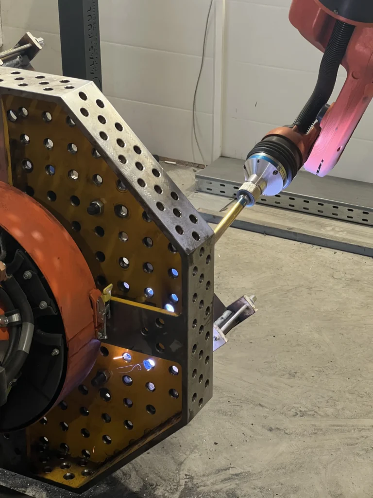 A robotic welding machine welds a steel part secured to a perforated welding table