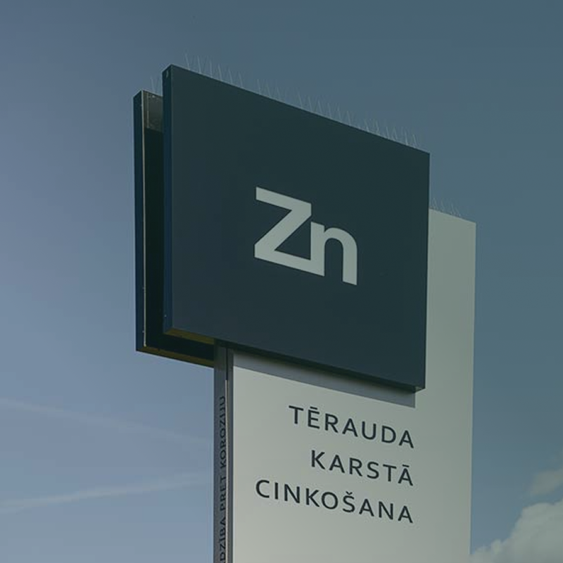 Zn Metals reklāmas zīme ar tekstu "Tērauda karstā cinkošana" pret zilām debesīm
